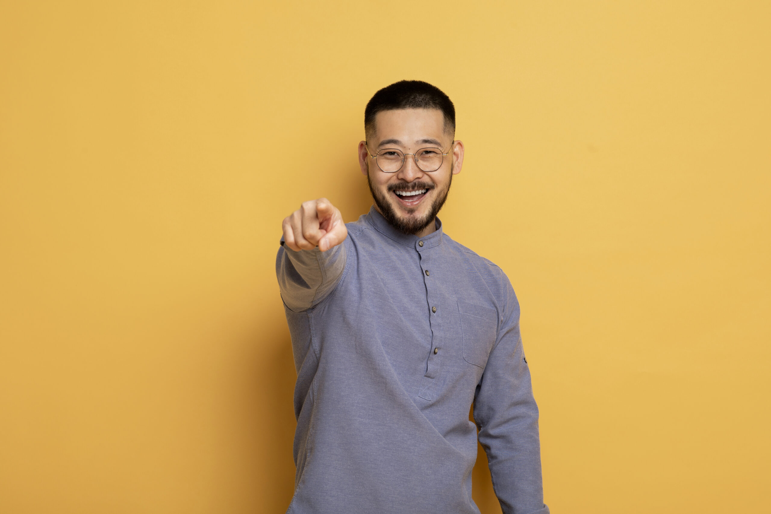 Portrait of young asian man pointing index finger at camera, cheerful millennial guy posing over yellow studio background, smiling male picking, choosing and indicating somebody, copy space