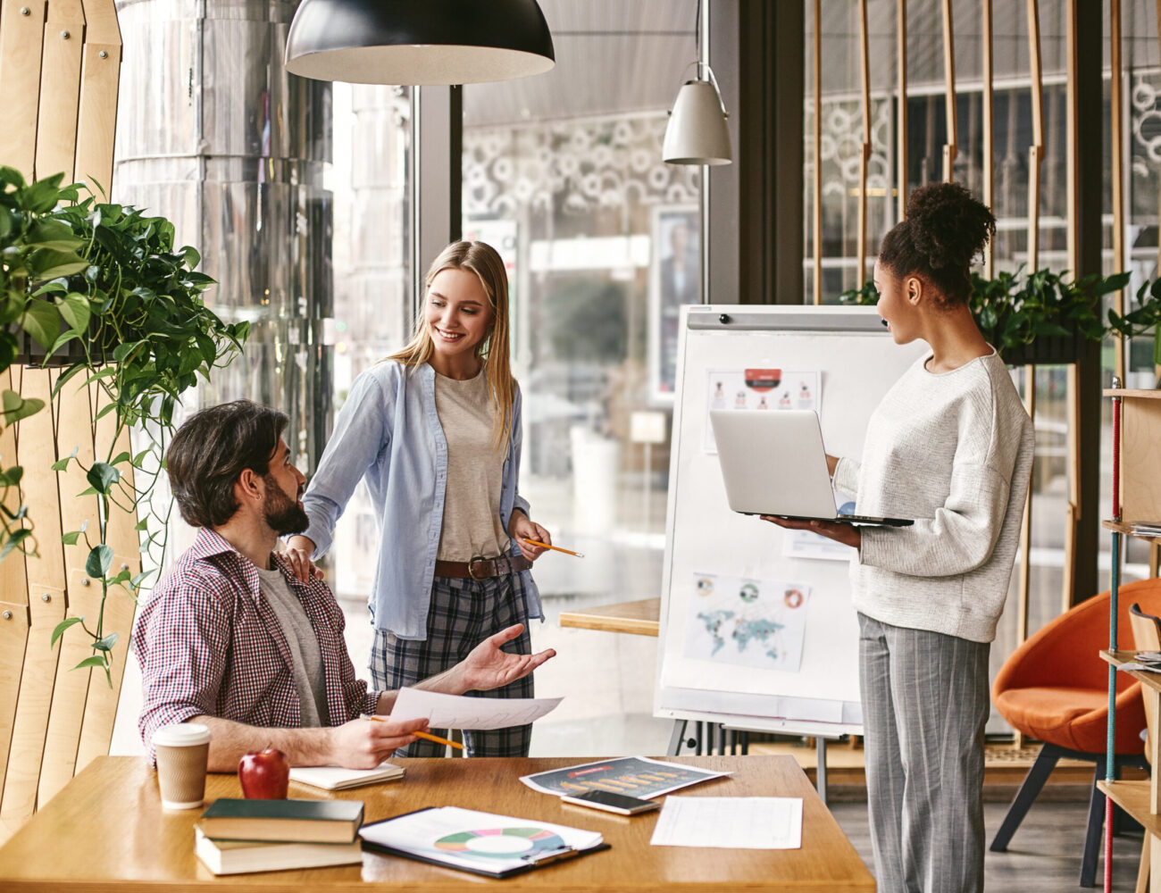 Attractive young woman standing near whiteboard with a laptop in her hands and looking to it. Another co-workers are talking to each other, looking with a smile. Concept of success