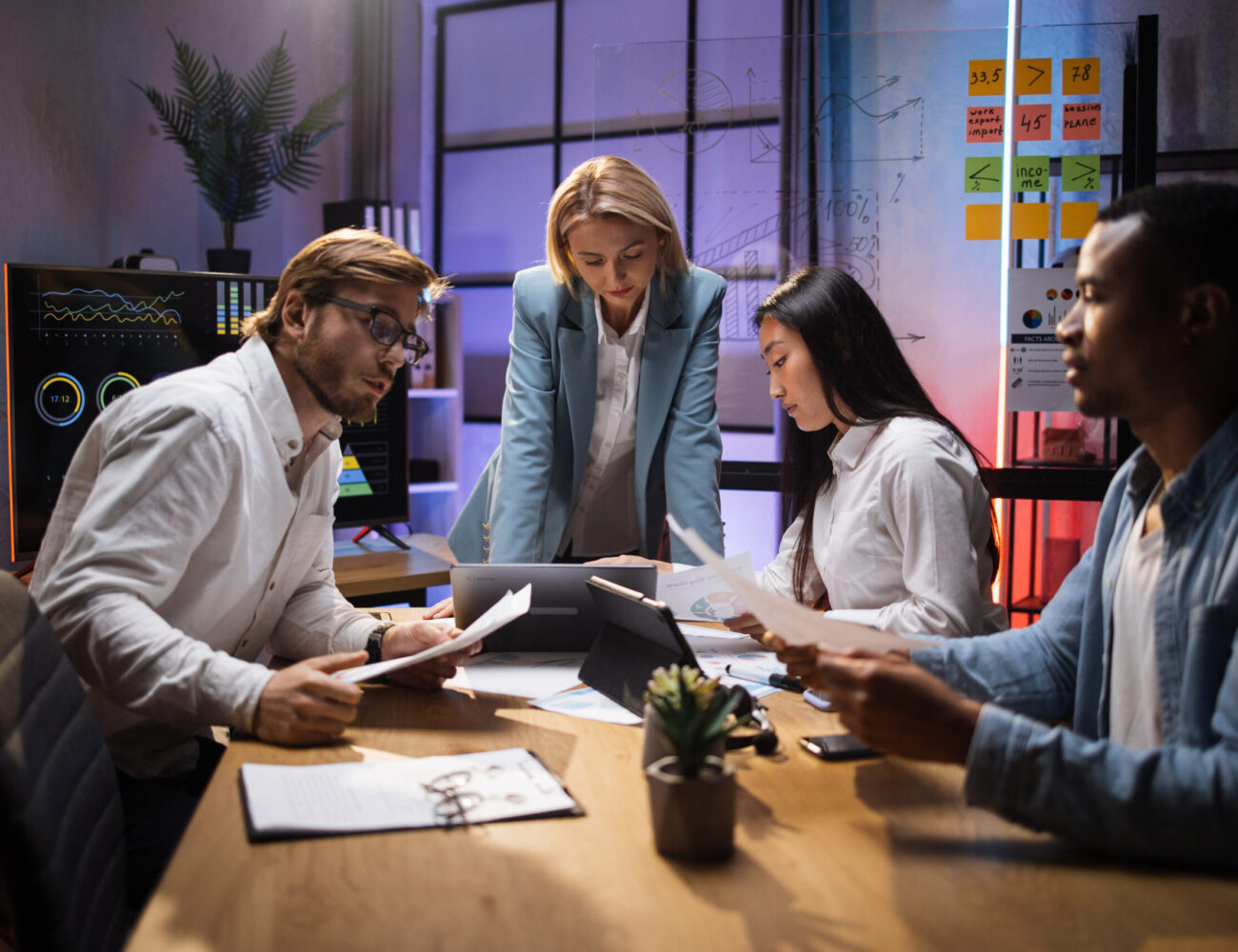 Female team leader in business clothes having briefing with multi ethnic colleagues at office room. Company workers sitting at desk, using modern gadgets and analysing financial statistics.