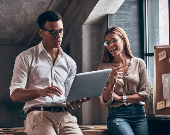 Two young coworkers in smart casual wear discussing business using laptop and smiling while working in the office